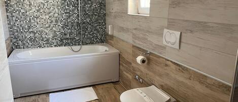 A modern bathroom with a mosaic tile shower wall, a freestanding bathtub, a toilet, and a window providing natural light.
