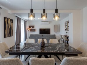 Un espace repas moderne avec une table en marbre, des chaises rembourrées, des luminaires suspendus et un téléviseur à écran plat fixé au mur.