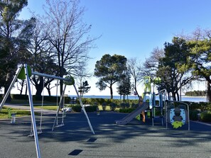 Un parque infantil con columpios y tobogán, rodeado de árboles y vista al mar.