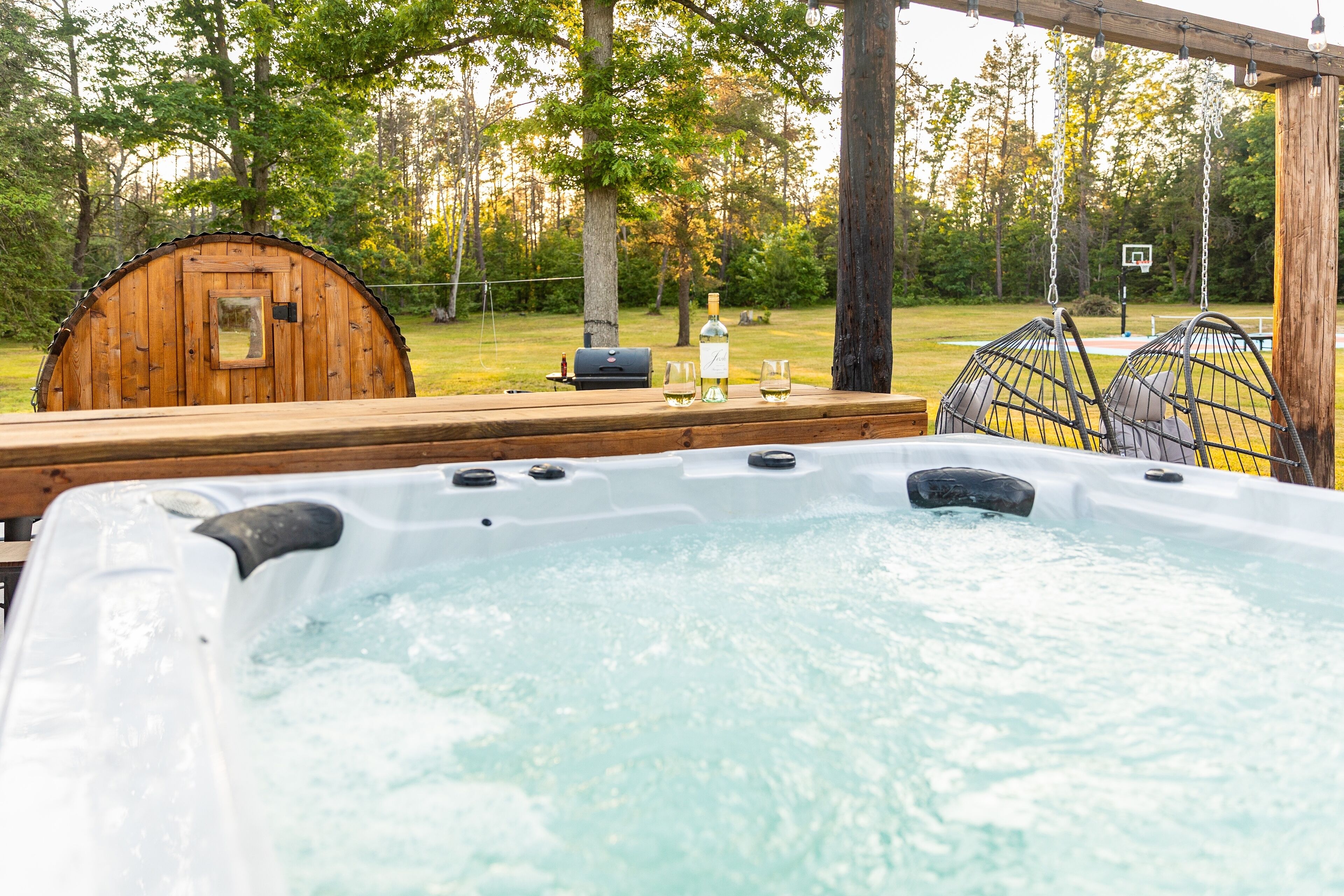 A hot tub with a wooden deck, a wooden barrel, and a basketball hoop in a wooded area.