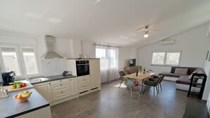 Eine moderne Küche mit weißen Schränken, ein Essbereich mit Holztisch und -stühlen sowie eine Wohnbereich mit Sofa und Fernseher.