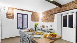 Un coin repas avec une table en bois dressée pour deux personnes, entourée de chaises blanches. Une fenêtre offre une vue sur l’extérieur, et les murs sont recouverts de pierres apparentes.