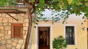 A stone building with a wooden door and shutters, a potted plant, and a vine with grapes. - Apartment Santic by Interhome (Krk/Čižići)