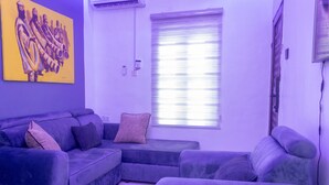 Un salón con un sofá morado, un cuadro grande en la pared y una ventana con persianas.
