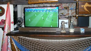 Eine Bar mit einem Fernseher, der ein Fußballspiel zeigt, einem Holzschild mit der Aufschrift „SOUVENIRS – einzigartige Handarbeit“ sowie verschiedenen Dekorationen, darunter ein kleines Schiffmodell und eine Flasche.