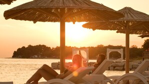 Two lounge chairs under thatched umbrellas on a pebbled beach with a sunset in the background. - Villa Linet 3 by Interhome (Poreč/Funtana)