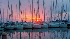 Un port esportiu amb diversos velers amarrats, que reflecteixen el sol ponent a l'aigua.