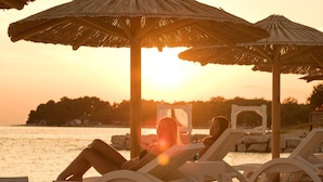 Eine Strandansicht mit zwei Liegestühlen unter Reetdachschirmen, mit Blick auf ein Gewässer, dessen Hintergrund von einem Sonnenuntergang bestimmt wird.