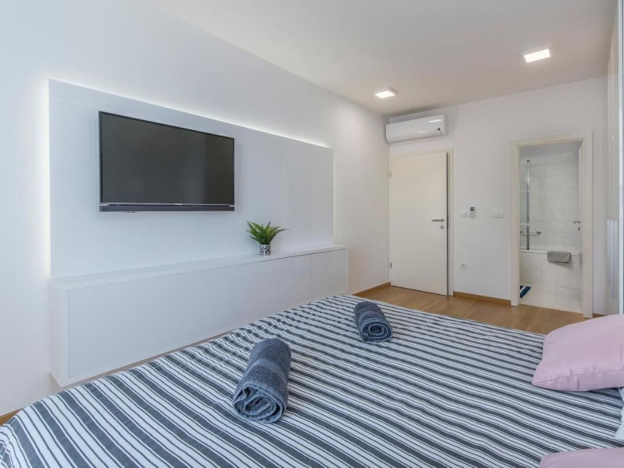 Ein modernes Hotelzimmer mit einem großen Bett, einem flachbildfernseher an der Wand, einer kleinen Topfpflanze und einer Duschkabine.