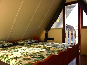 A bedroom with a sloped ceiling, a bed with a floral bedspread, a red bedside lamp, and a view of a house through a window. - Haus Reiher am Lauwersmeer by Interhome (Anjum)