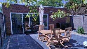 Eine Terrasse mit Holztisch und -stühlen, ein Glastür-Eingang und eine Ziegelwand.