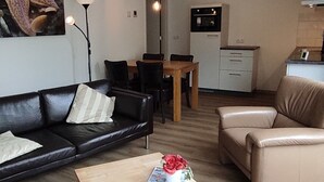 Ein Wohnzimmer mit einem schwarzen Ledersofa, einem beigefarbenen Sessel, einem Couchtisch aus Holz mit einer Zeitschrift und einer Blumenvase, ein Essbereich mit Holztisch und Stühlen sowie eine Küchenzeile mit eingebautem Ofen und Kühlschrank.