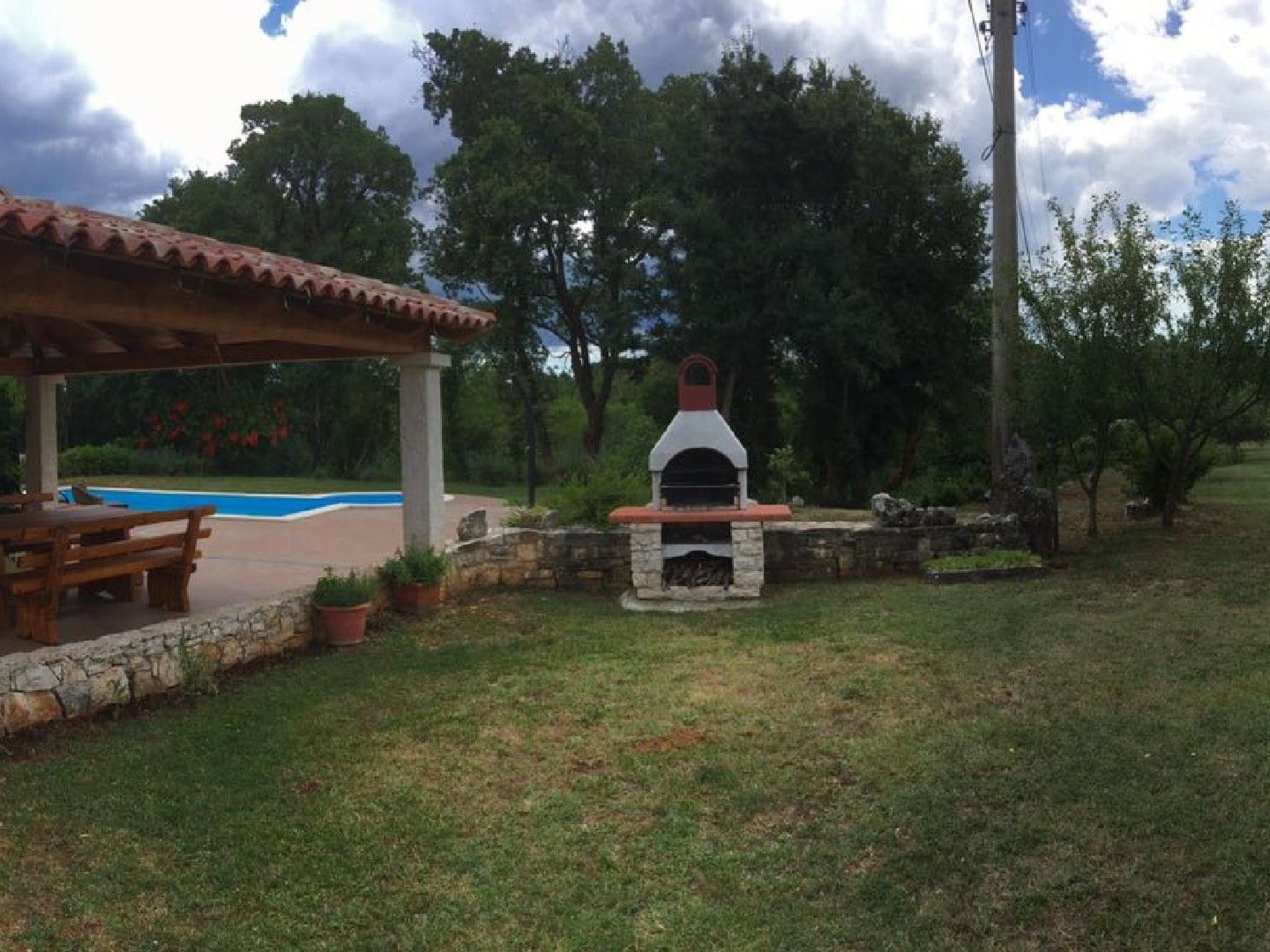 Un área de barbacoa al aire libre con horno de piedra, una zona de estar cubierta y una piscina al fondo.