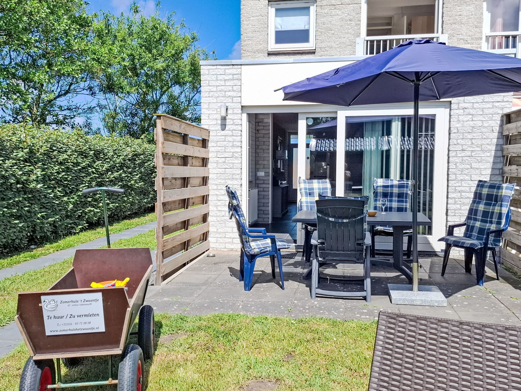 An outdoor patio with a table and chairs, a blue umbrella, and a wooden cart with a sign.