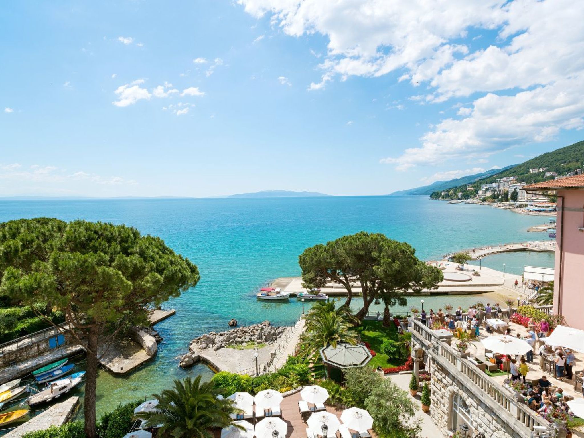 A coastal resort with a marina, outdoor seating, and a view of the sea and distant hills.