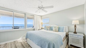 A bedroom with a bed, a ceiling fan, a nightstand, a lamp, and a view of the ocean. - Oceanfront 2BR Condo with Balcony & Beach Access (Fernandina Beach)