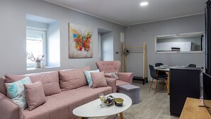 Un salon moderne avec un canapé rose, une table basse, un coin repas avec une table et des chaises, un coin cuisine et un tableau coloré représentant un papillon accroché au mur.