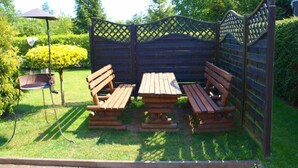 Un espace extérieur aménagé avec des sièges en bois, comprenant une table et des bancs, entouré d’une clôture en treillis et de verdure.
