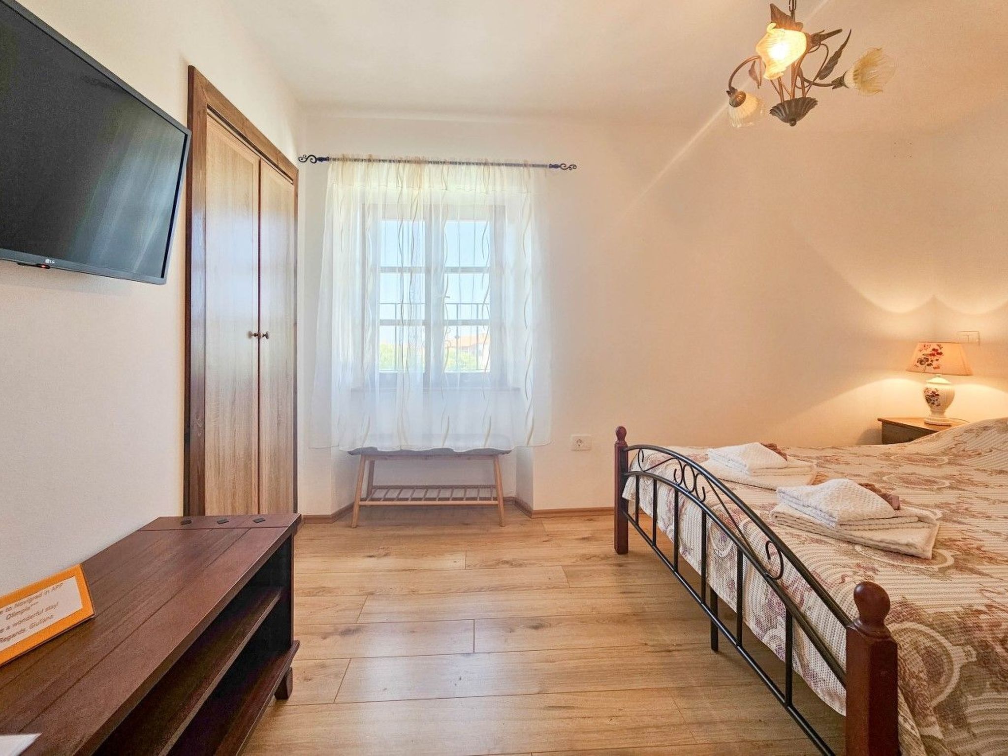 Una camera da letto con un letto, una TV a muro, una panca di legno e una finestra con tende leggere.