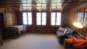 A cozy room with a wooden ceiling, a sofa, a table with chairs, and large windows.