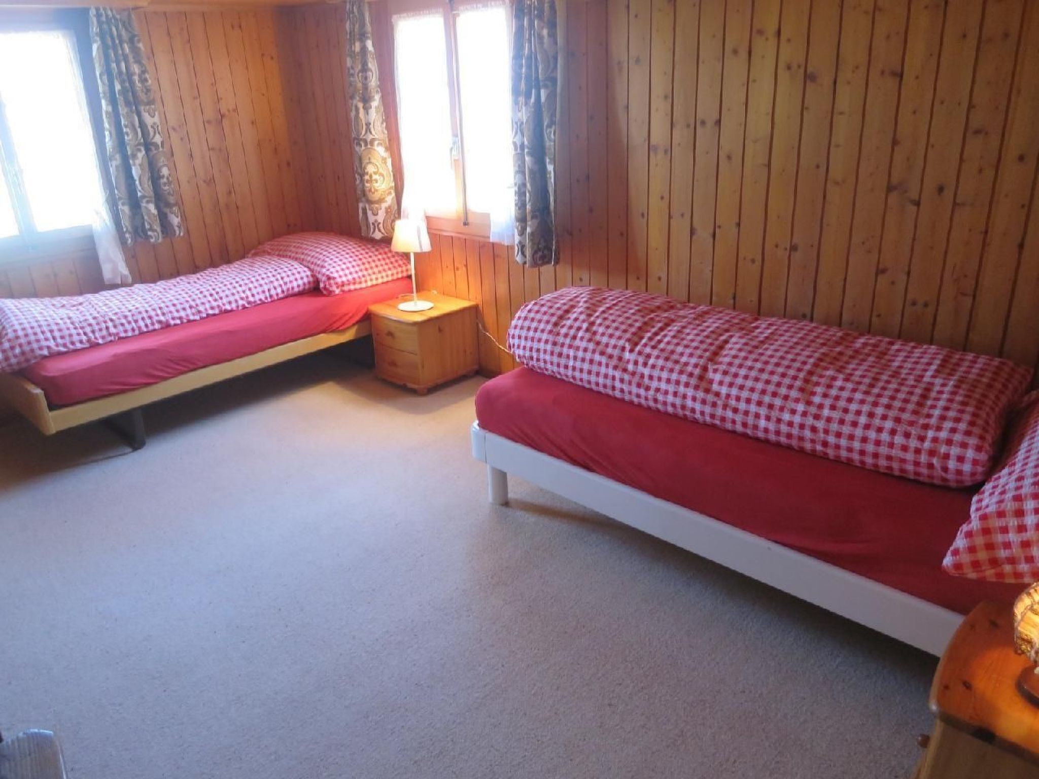 Ein Zimmer mit zwei Betten, jeweils mit roter Bettwäsche und karierten Kissen. Dazwischen steht ein Nachttisch aus Holz mit einer Lampe. Die Wände sind aus Holz und der Boden ist mit Teppich ausgelegt.