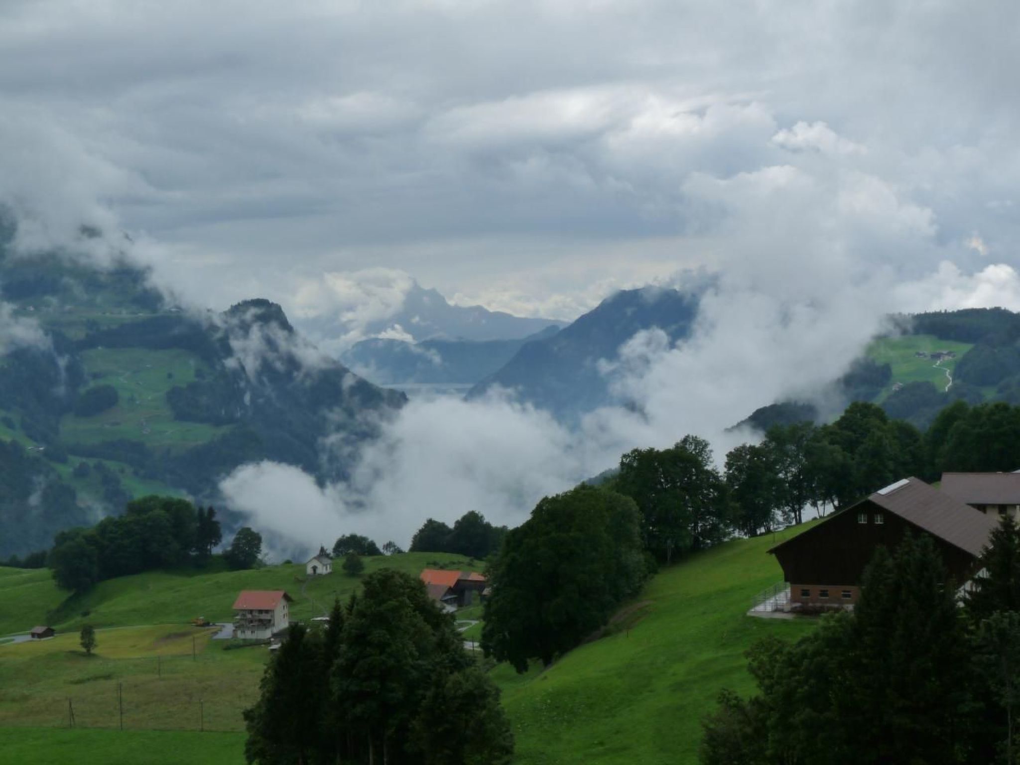 Ländliche Landschaft mit Häusern an einem Hügel, umgeben von Bergen und Wolken.