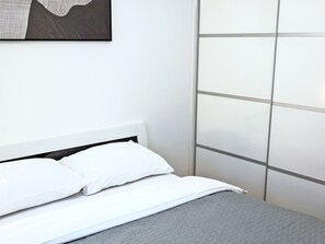 Ein ordentlich bezogenes Bett mit weißer Bettwäsche und einer grauen Tagesdecke, ein weißer Kopfteil und eine Wand mit Rillenmuster.
