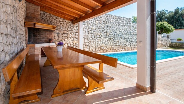 Une terrasse extérieure avec une table et des bancs en bois, un mur de pierre et une piscine en arrière-plan.
