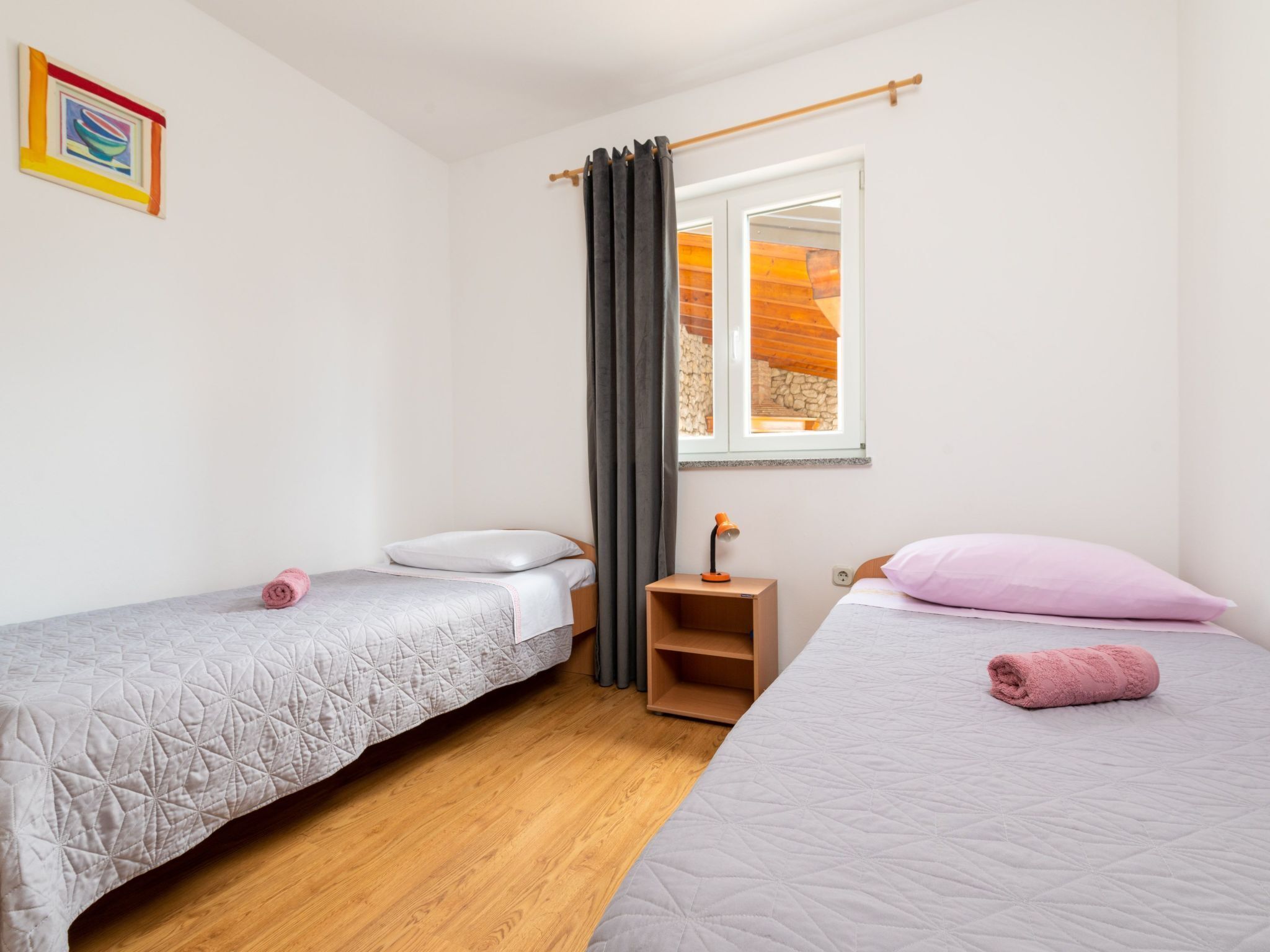 Un dormitorio con dos camas individuales, suelo de madera, una ventana que da a un muro de piedra y una mesita de noche con una lámpara.