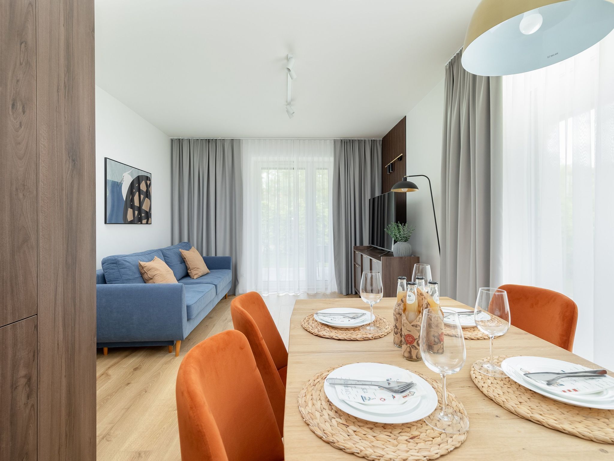 Een moderne woonkamer met een blauwe bank, een eettafel gedekt met borden, glazen en bestek, en een groot raam met gordijnen.