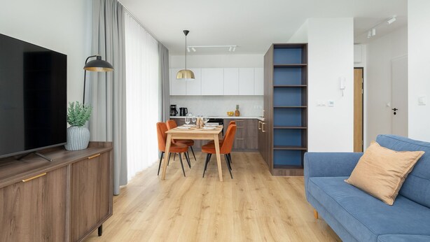 Una sala moderna con un sofá azul, un comedor con mesa y sillas de madera, una cocina al fondo y un televisor a la izquierda.