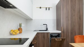 Una cocina moderna con encimera blanca, armarios de madera, horno empotrado y un pequeño comedor con mesa de madera y silla naranja.