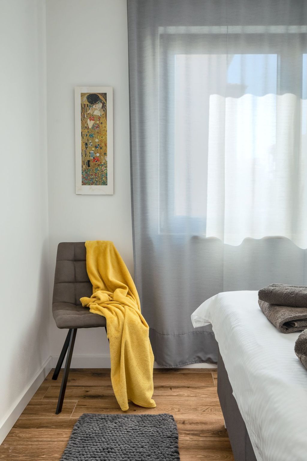 A bedroom with a bed, a chair with a yellow blanket, a gray rug, a gray curtain, and a framed picture on the wall.