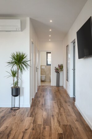 Ein Flur mit Holzboden, einer Zimmerpflanze, einem an der Wand montierten Fernseher und einem im Hintergrund erkennbaren Badezimmer.