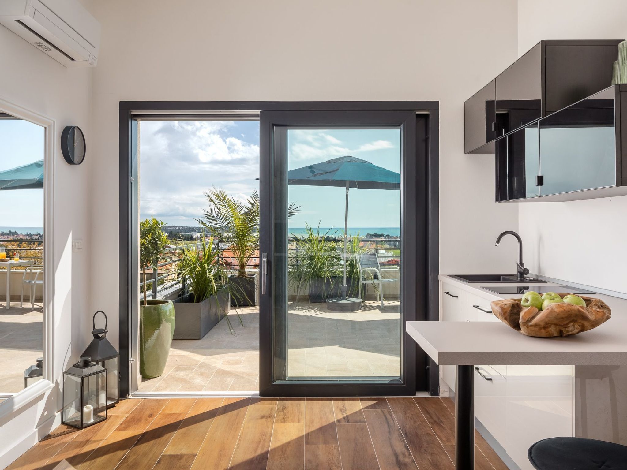 A modern kitchen with a view of the sea, a dining area with a table and chairs, and a balcony with plants and an umbrella.