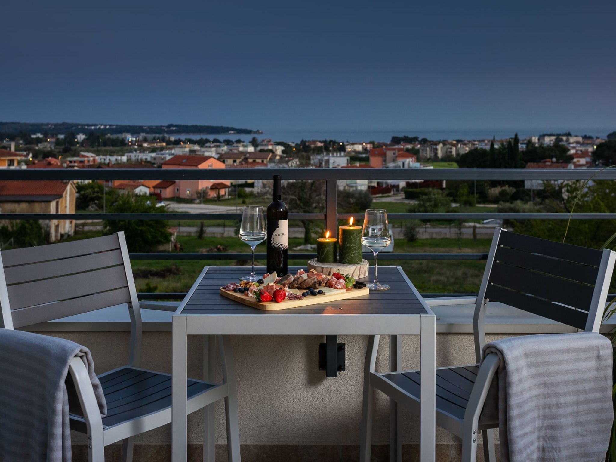 A table set with a wine bottle, glasses, candles, and a charcuterie board on a balcony overlooking a coastal town.