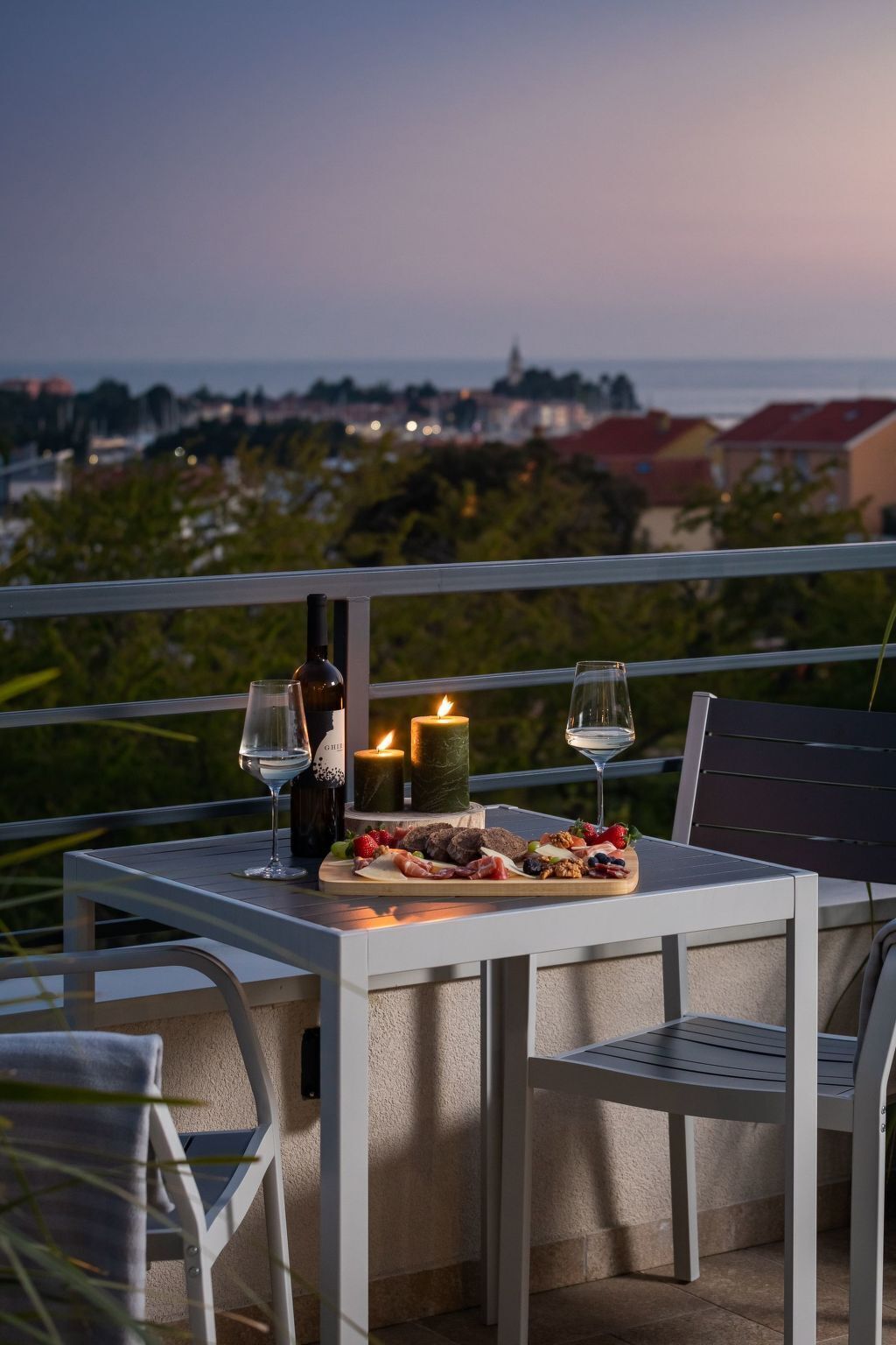 A balcony with a table set for a meal, including a bottle of wine, glasses, a candle, and a fruit platter.