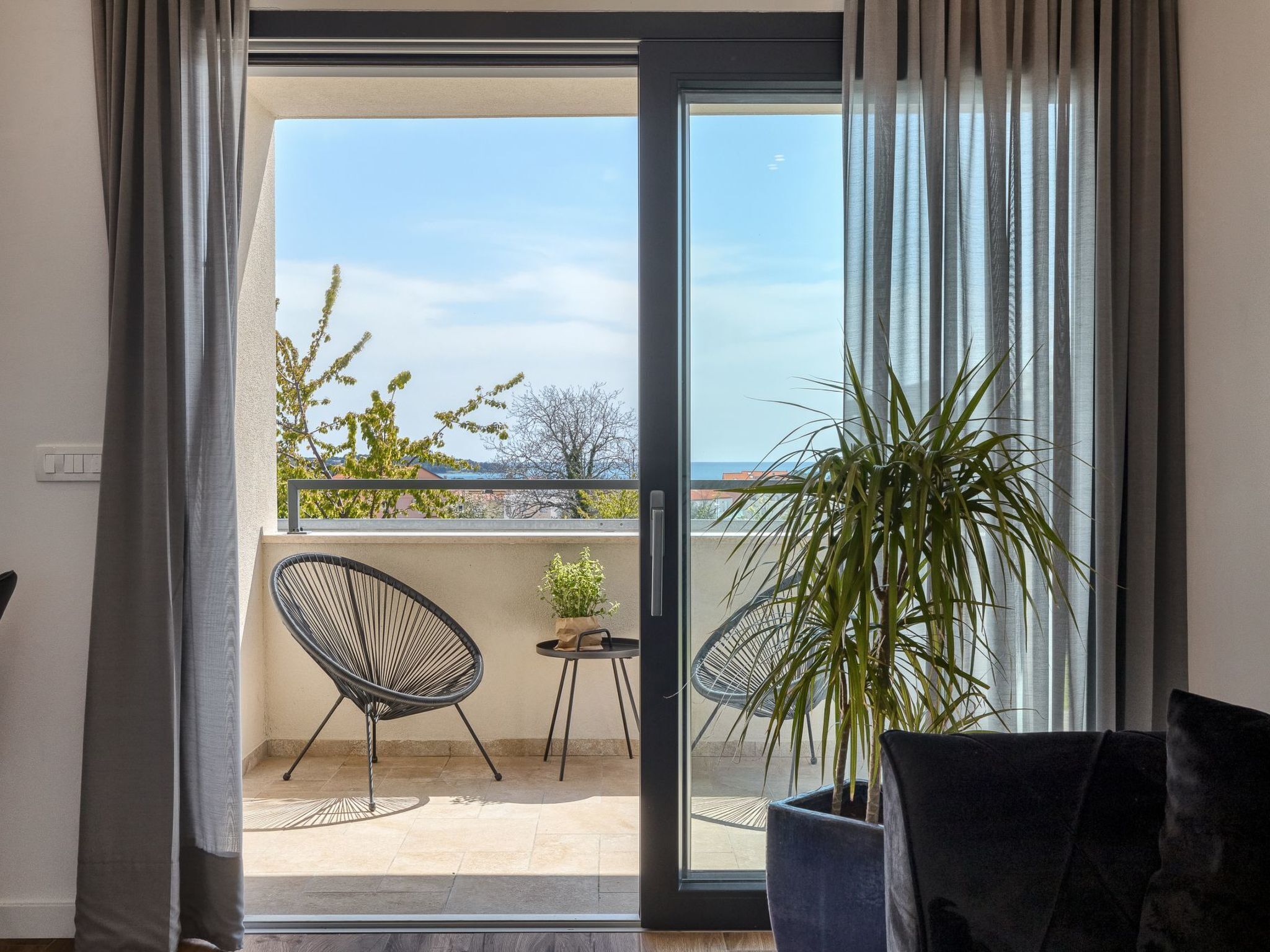 A balcony with a view of the sky and trees, a chair, a small table, and a potted plant.