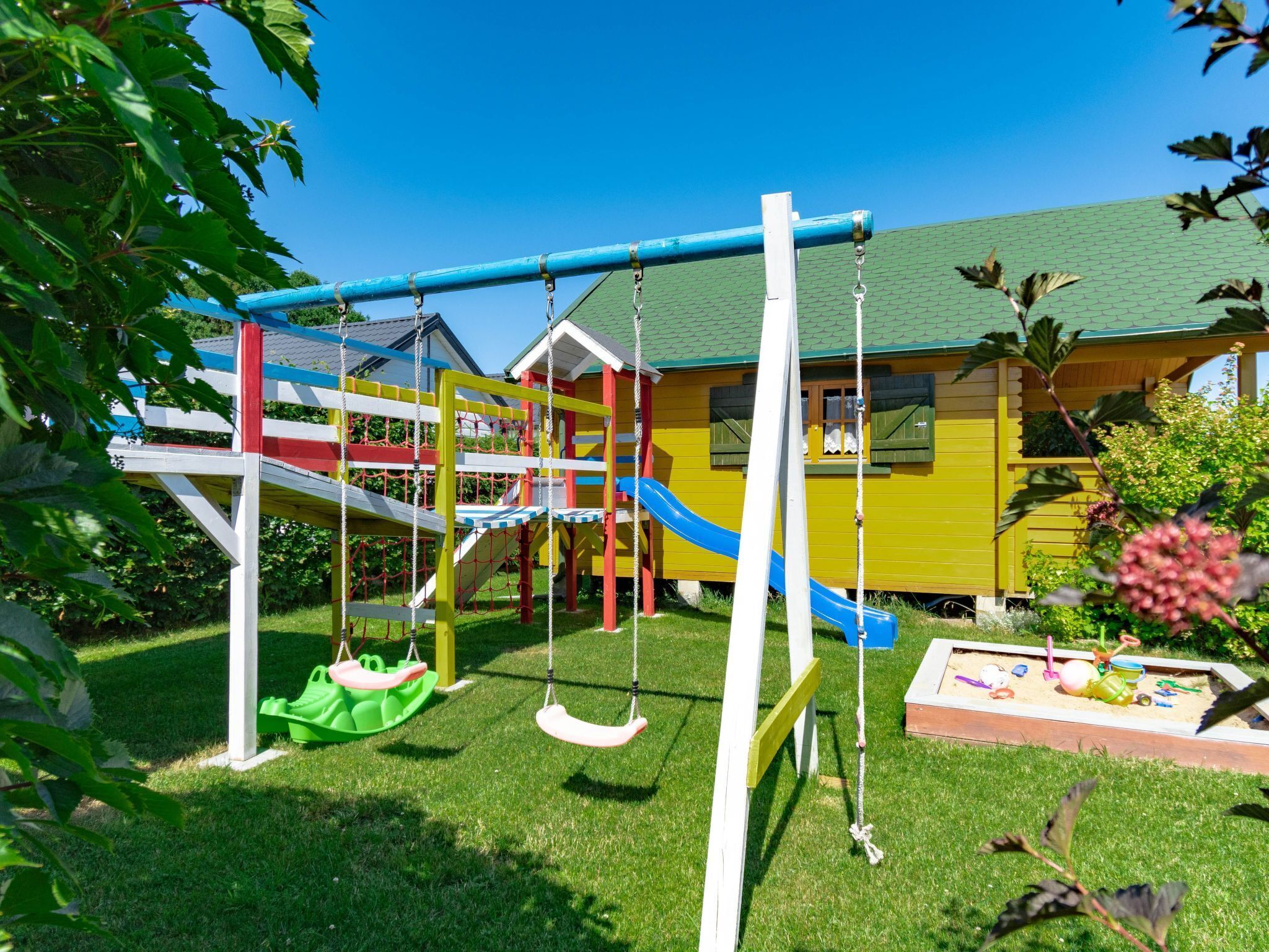 Ein Spielplatz mit Schaukeln und Rutschen, einer grünen Wiese und ein gelbes Gebäude mit einem grünen Dach im Hintergrund.