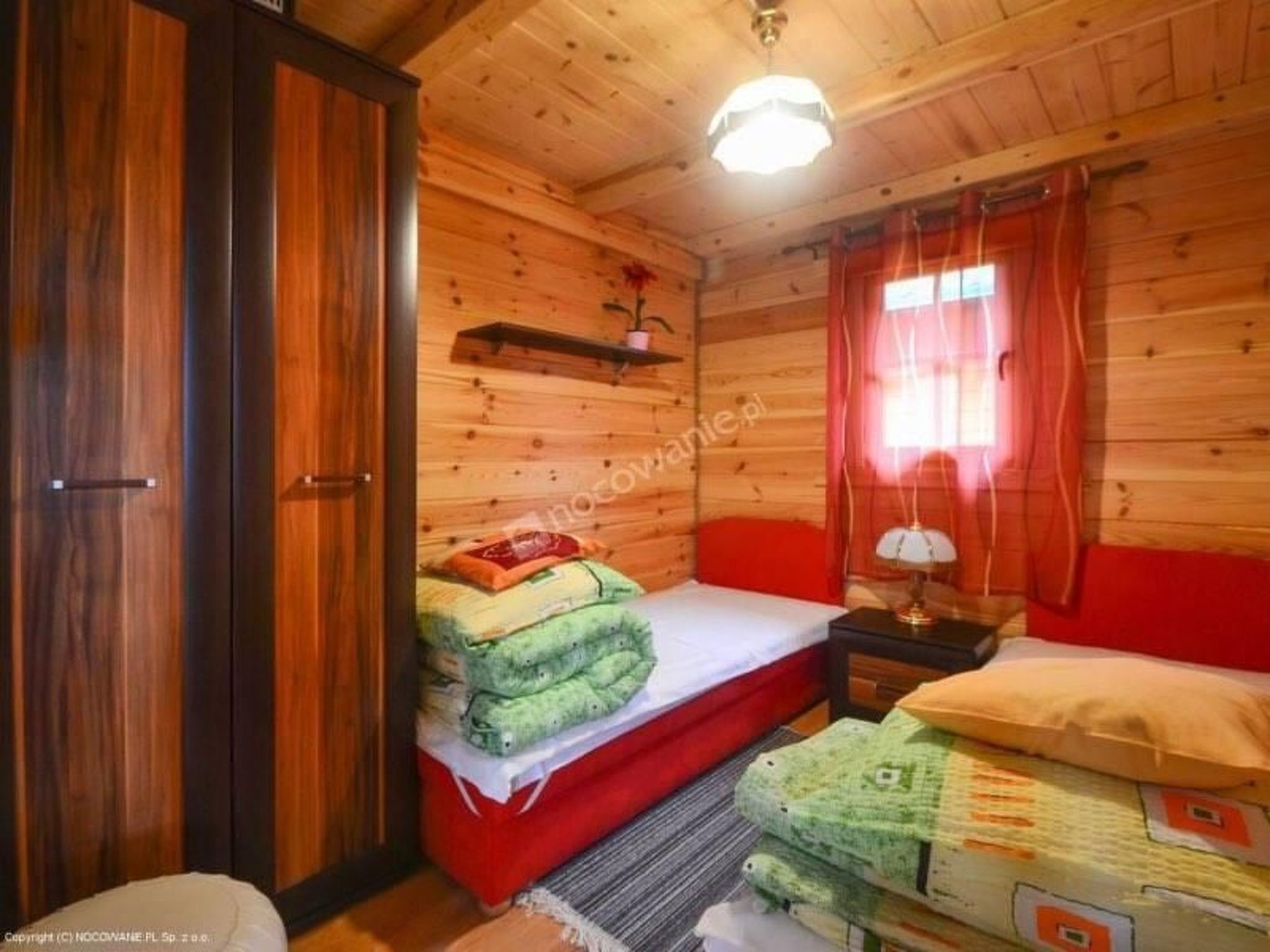 Une pièce en bois avec un lit, une table de chevet, une lampe et une fenêtre ornée de rideaux rouges.