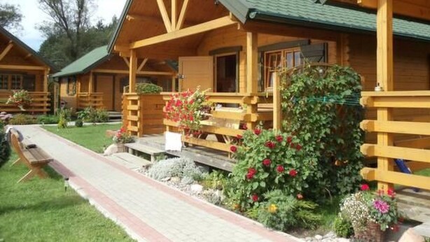 Une cabane en bois avec un toit vert, une véranda dotée d’une balustrade en bois et un jardin agrémenté de fleurs et d’arbustes.