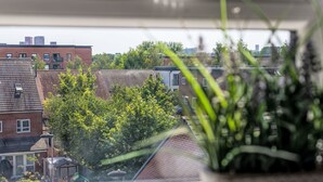 Blick durch ein Fenster mit einem Blumentopf im Vordergrund, ein Backsteinhaus und ein Baum im Hintergrund.