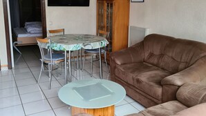 A living room with a brown sofa, a glass-top coffee table, a dining table with chairs, a TV mounted on the wall, and a cabinet with shelves. - Vacation home House by Interhome (Bredene)