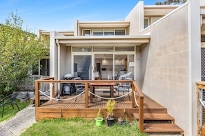 A modern house with a deck, outdoor seating, and a kitchen visible through glass doors. - Coastal Nook (Lorne)