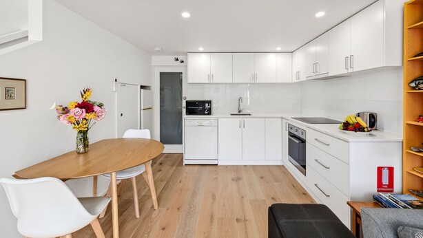 A modern kitchen with white cabinets, a dishwasher, and a microwave. There is a dining table with chairs and a vase of flowers. - Coastal Nook (Lorne)