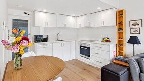 A modern kitchen with white cabinets, a microwave, and a sink. There is a dining table with a vase of flowers. - Coastal Nook (Lorne)