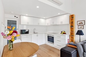 A modern kitchen with white cabinets, a microwave, and a sink. There is a dining table with a vase of flowers. - Coastal Nook (Lorne)
