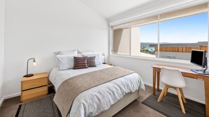 A bedroom with a bed, bedside table, desk, chair, and a window with a view. - Coastal Nook (Lorne)