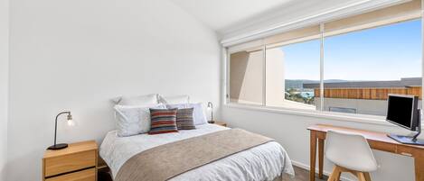 A bedroom with a bed, bedside table, desk, chair, and a window with a view.