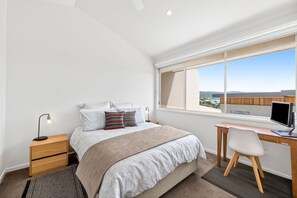 A bedroom with a bed, bedside table, desk, chair, and a window with a view. - Coastal Nook (Lorne)